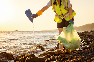 Volunteer in orange vest picking up plastic waste and debris from beach shore.