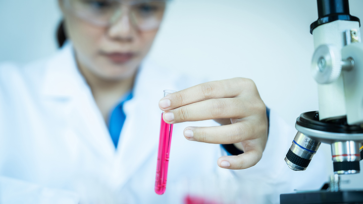 A woman in a lab coat holding a test tube with a pink liquid in it. 
