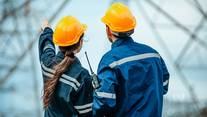 Workers examine and discuss power lines while wearing safety gear at an electrical substation