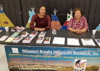 Missour Breaks table at event, with two tribe members looking at the camera