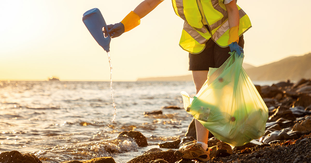 Volunteer in orange vest picking up plastic waste and debris from beach shore.