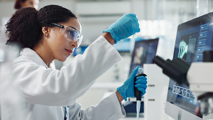 Scientist in safety goggles and gloves, runs an experiment in front of a computer screen