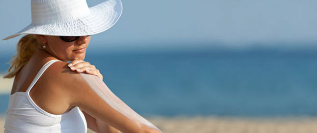 A woman wearing protective clothing at the beach putting sunscreen on her arm