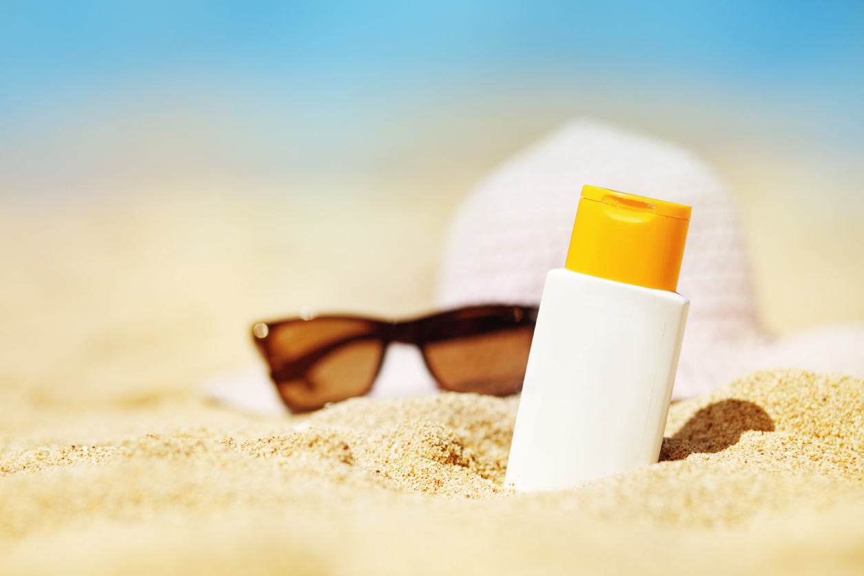 closeup of bottle of sunscreen slightly buried in sand with sunglasses and hat blurred in the background