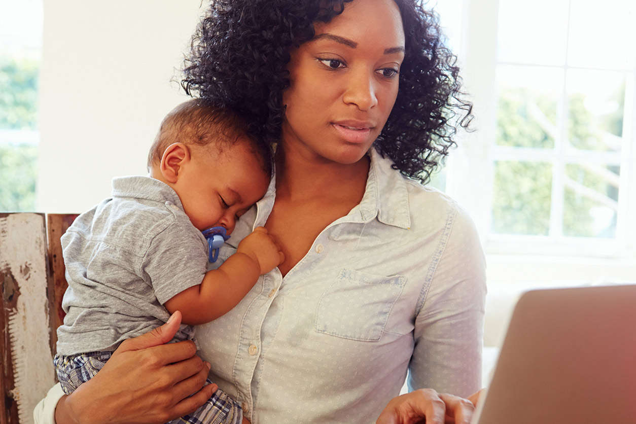 woman holding a sleeping baby while looking at a laptop